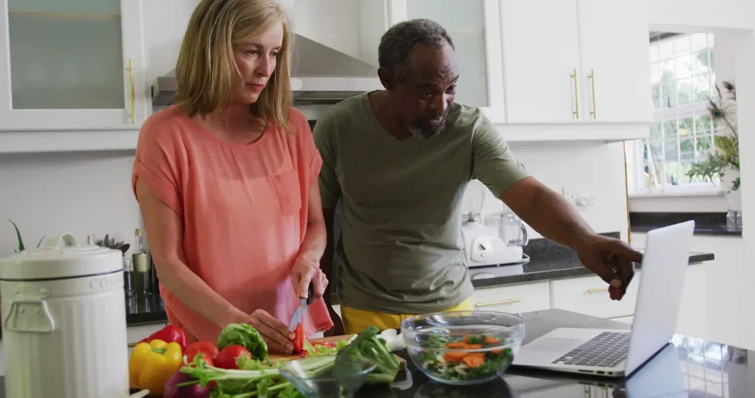 Diverse senior couple preparing food in kitchen following recipe on laptop - Image